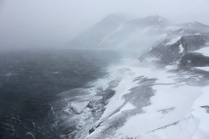 Snow-covered rocky coastline with strong winds and rough sea waves in Antarctica landscape under cloudy sky.