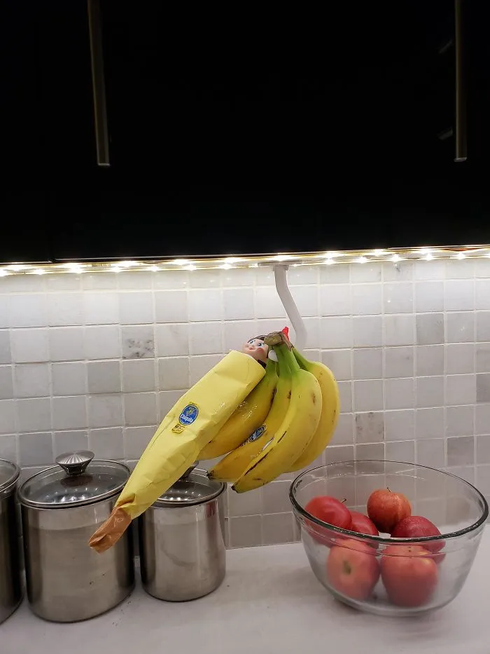 Elf On The Shelf hiding in a banana peel sleeve among bananas in the kitchen, showcasing a playful mischief idea.