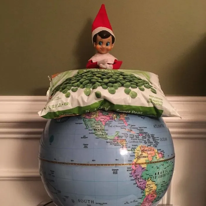 Elf On The Shelf sitting on a globe resting on frozen peas bag, creatively representing world peace in a playful holiday setup.