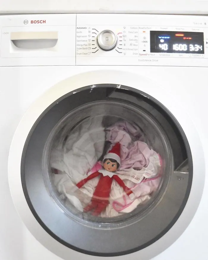 Elf On The Shelf idea with the elf inside a washing machine drum surrounded by clothes for mischievous holiday fun.