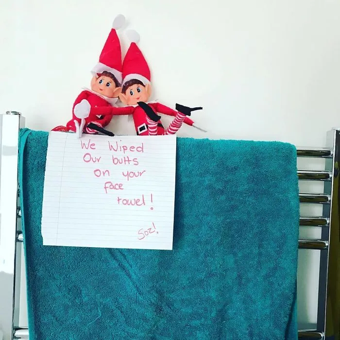 Two Elf On The Shelf dolls on a towel rack with a mischievous note on a face towel prank idea.