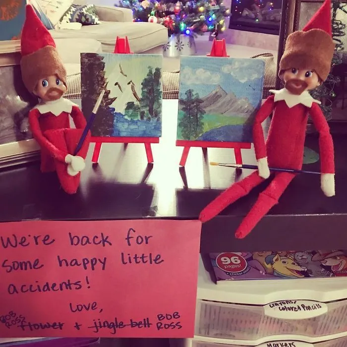 Two Elf On The Shelf dolls dressed as Bob Ross, painting miniature landscapes with a sign about happy little accidents.