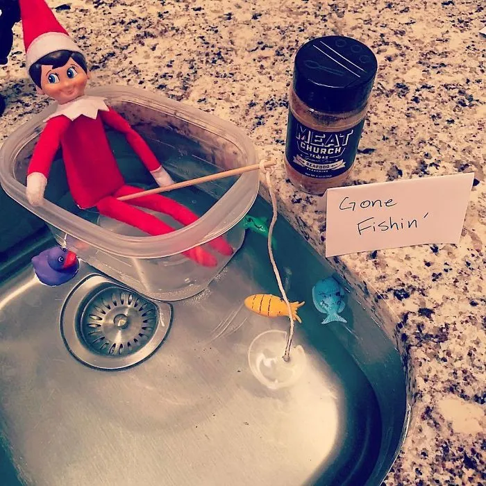 Elf On The Shelf idea with an elf fishing in a plastic boat in the kitchen sink surrounded by toy fish and a gone fishin sign.
