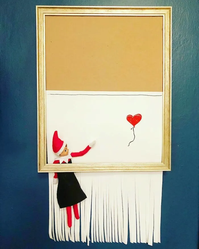 Elf On The Shelf idea with elf in a frame reaching for a red heart balloon amid shredded paper below.