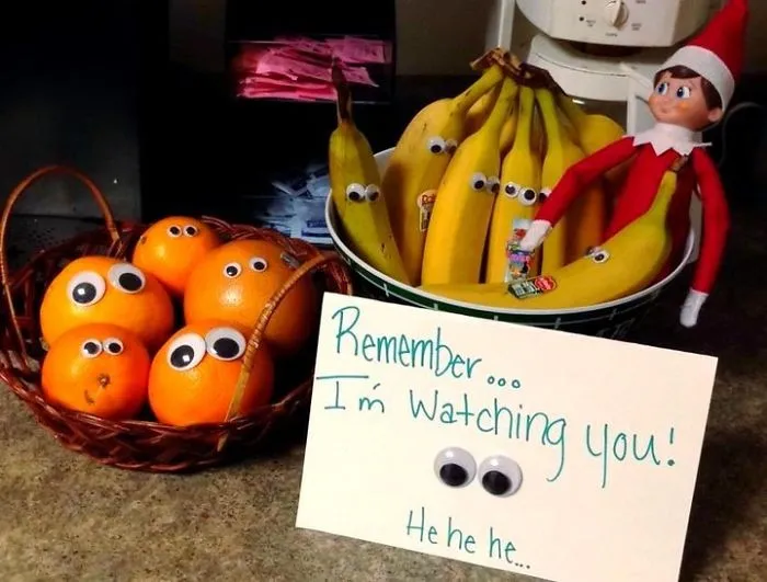 Elf On The Shelf with googly-eyed bananas and oranges, holding a sign saying Remember I'm watching you in a kitchen setting