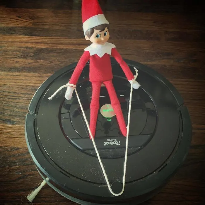 Elf On The Shelf skiing on a Roomba vacuum holding strings as ski poles for a creative Elf On The Shelf idea.