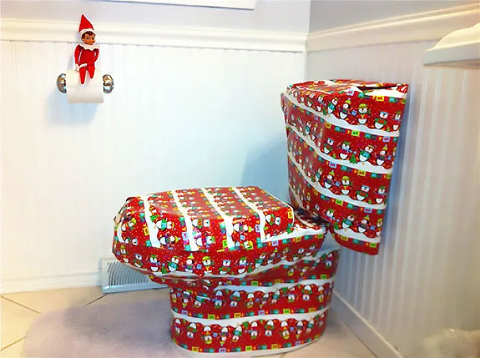 Elf on the Shelf perched on toilet paper holder with toilet fully wrapped in festive holiday gift wrap prank.