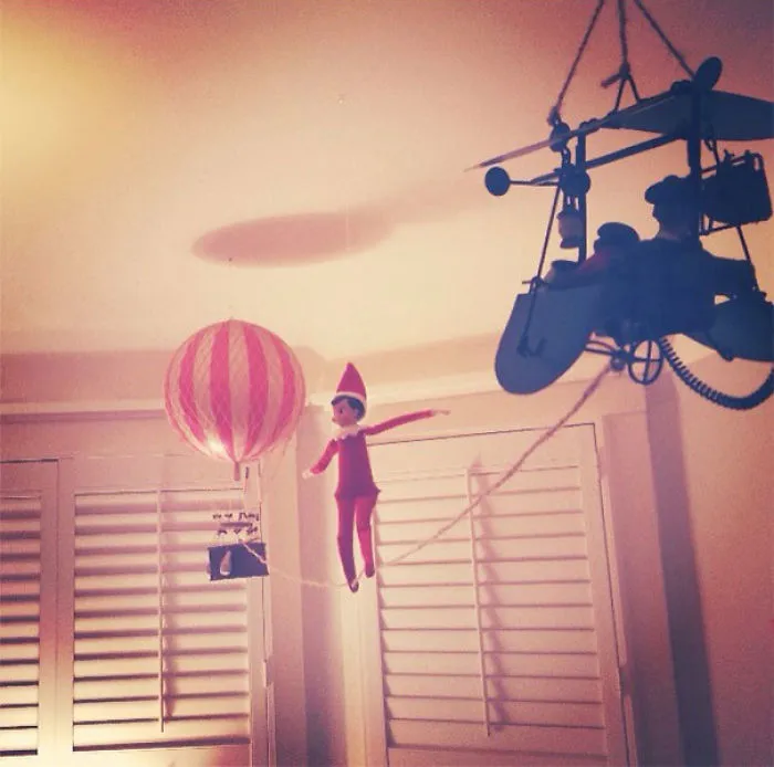 Elf On The Shelf balancing on a tightrope between a hot air balloon and airplane, creating an adventurous mischief scene.
