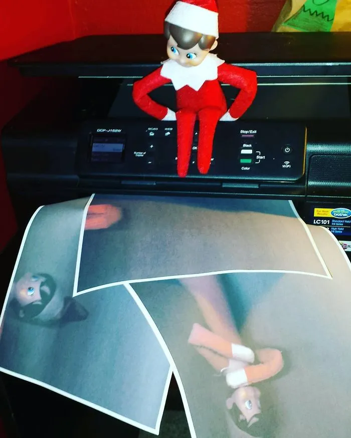 Elf On The Shelf sitting on a printer, creating enlarged copies of its own face on printed pages as a fun mischief idea.