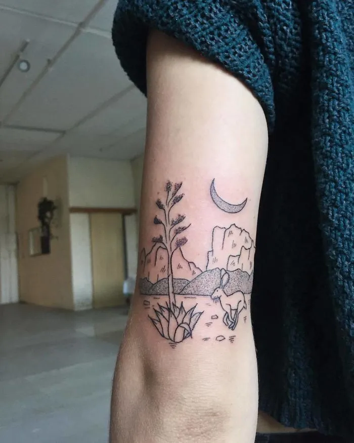 Arm with tattoo of desert scene, featuring a plant, rabbit, mountains, and crescent moon. Perfect for ink lovers.