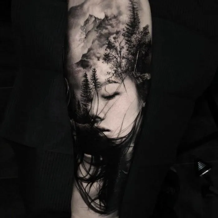 Dreamy landscape tattoo on arm featuring a woman's face with trees and mountains for ink lovers.