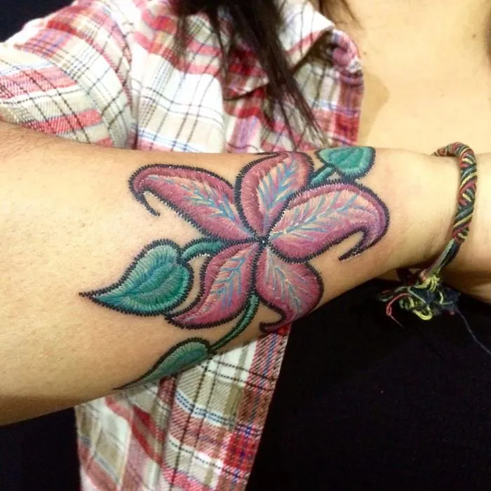Colorful flower embroidery tattoo on a person's forearm wearing a plaid shirt and bracelet.