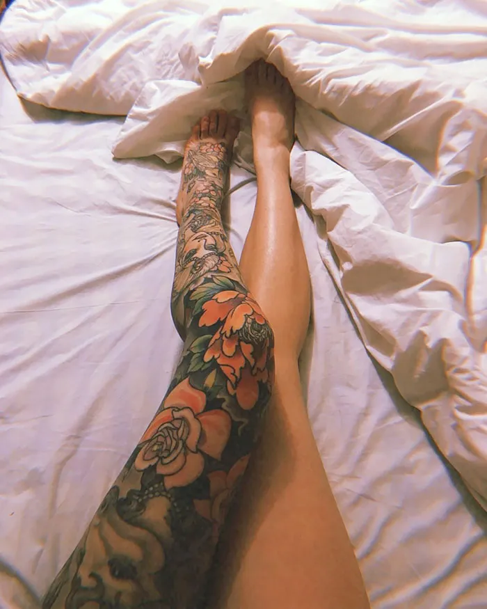 Colorful floral leg sleeve tattoo on a person lying on white sheets.