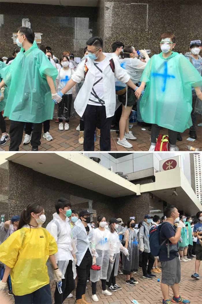 Voluntarios con ponchos marcados con una cruz azul, mascarillas y gafas, formando una pared humana para proteger la zona de primeros auxilios