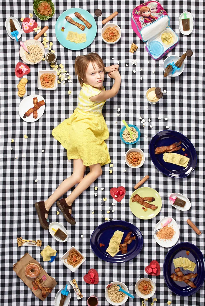 A young girl lies surrounded by her week's food, including bacon, pasta, and cookies, on a black and white checkered sheet.