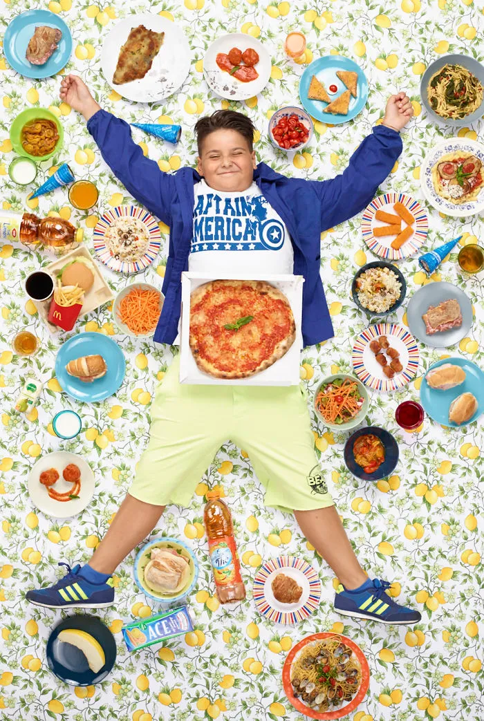 A boy smiles surrounded by a week's worth of food, including pizza, chicken, and fruit, on a lemon-patterned sheet.