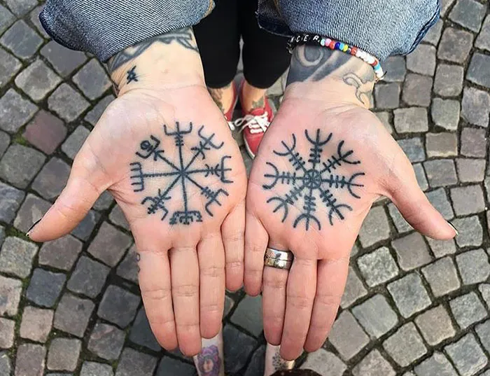Hands showing intricate palm tattoos with black ink designs on cobblestone background.