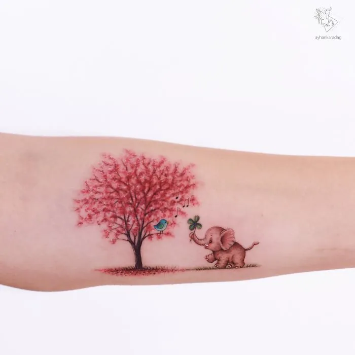 Tattoo design featuring an elephant with a clover, a vibrant tree, and a bluebird on human skin.