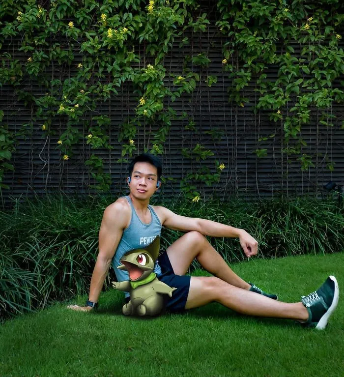 A man sitting on grass next to a Larvitar Pokémon illustration, surrounded by green foliage.