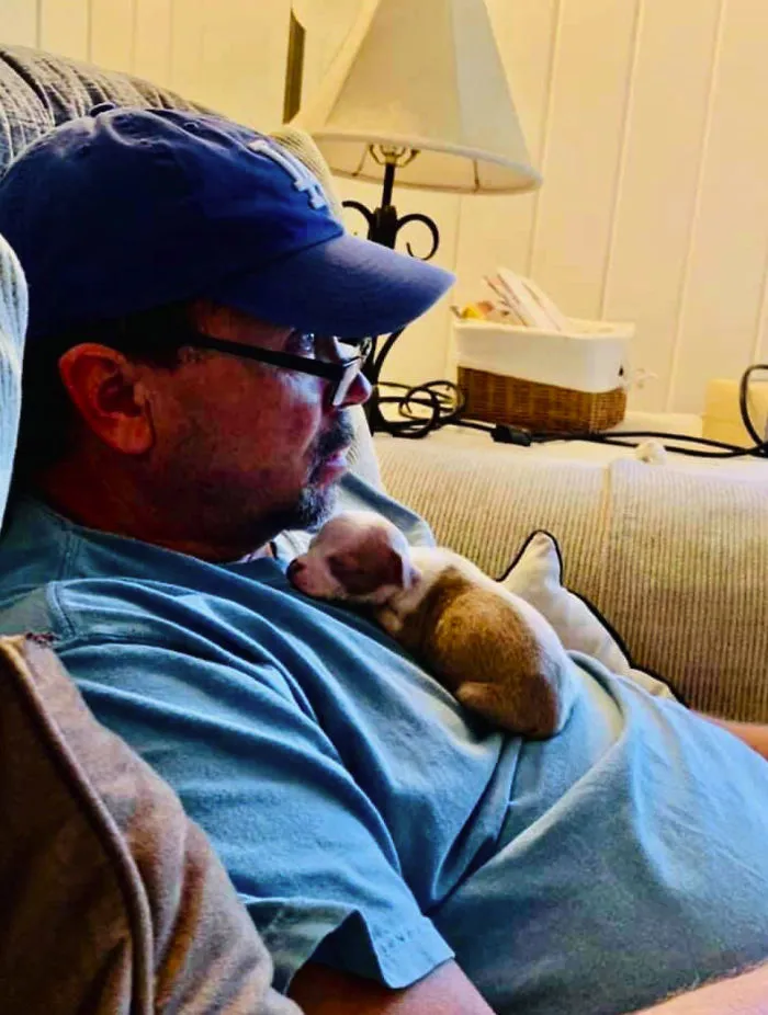 Man wearing glasses and blue cap resting on couch with a small rescue puppy sleeping on his chest, showing wholesome pet bond.