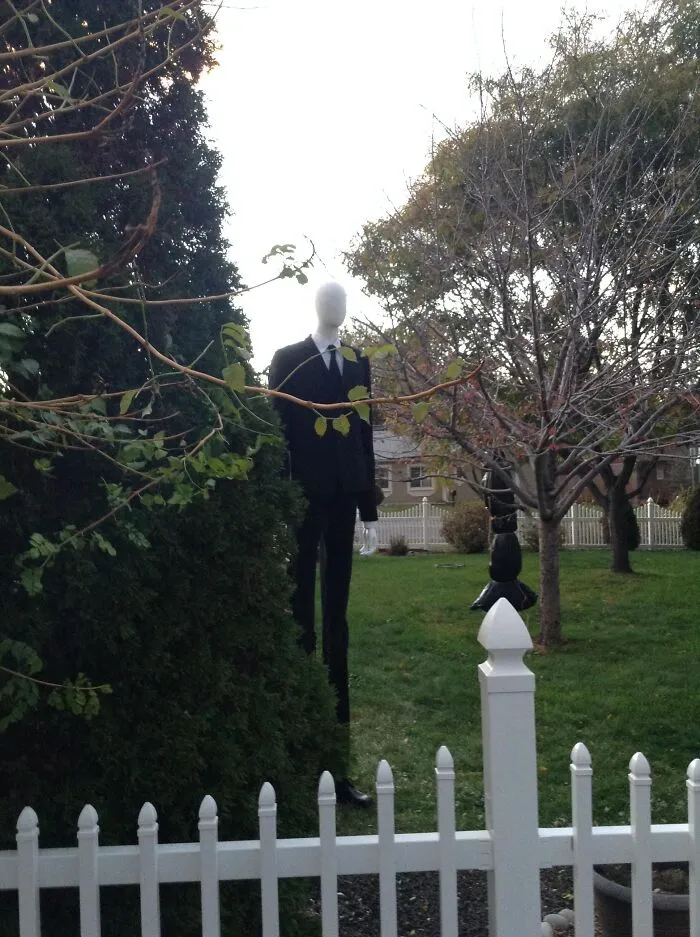 Slender Halloween decoration wearing a black suit and tie standing behind a tree in a yard with a white picket fence.