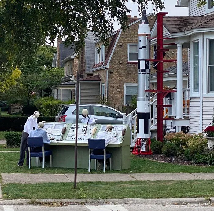 Halloween decorations featuring skeleton astronauts operating a vintage control panel next to a rocket in a suburban yard.