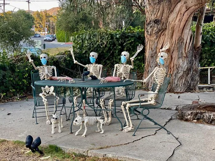 Four skeleton Halloween decorations wearing masks seated around a table with two skeleton dogs outdoors.