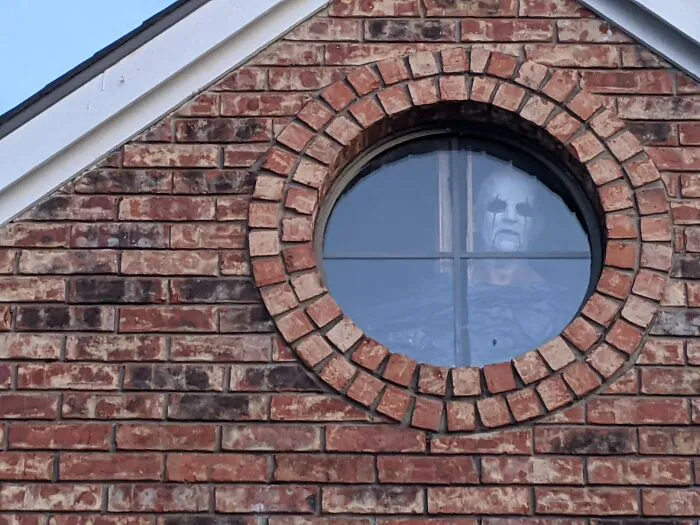 Creepy Halloween decoration of a ghostly figure with face paint seen through a round window in a brick house.