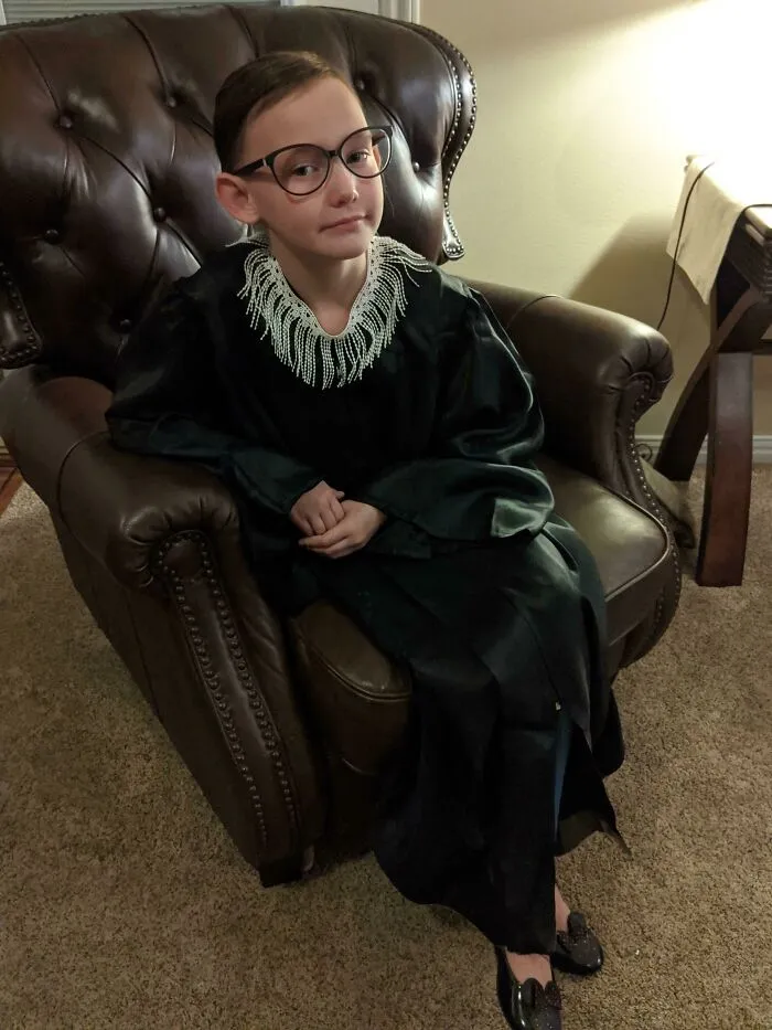 A young girl sitting in an armchair dressed in a Ruth Bader Ginsburg Halloween costume.