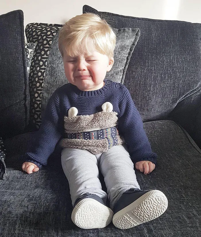 Toddler with closed eyes sitting on couch crying, illustrating kids are not born smart in a funny way.