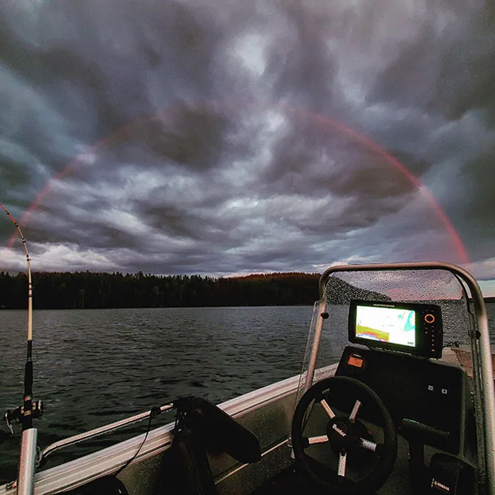 Boat under a rare rainbow amidst cloudy skies over a lake, revealing an unseen side of nature's beauty.