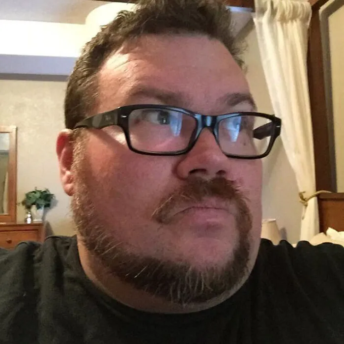 Man with glasses sporting a unique monkey tail beard style in a casual indoor setting.