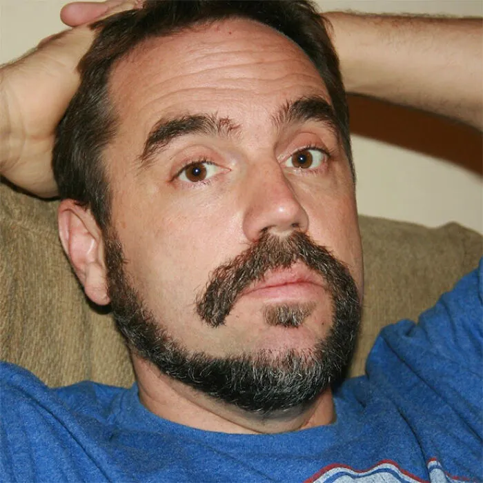 Man with a 'Monkey Tail' beard style, wearing a blue shirt, laying back on a couch.