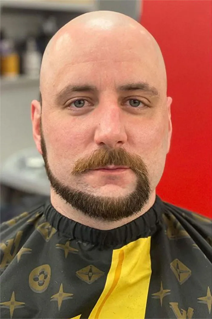 Man with a monkey tail beard style, bald head, wearing a patterned barber cape.