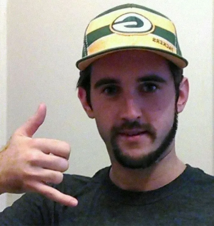 Man with a monkey tail beard and cap, gesturing a shaka sign.