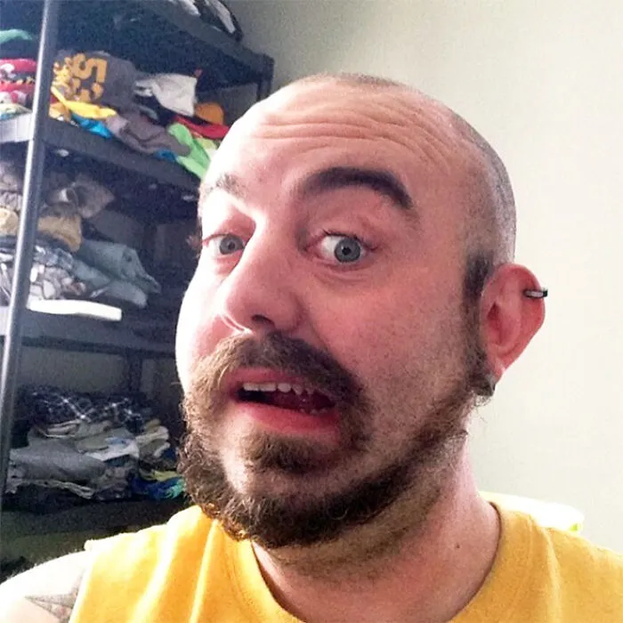 Man with a monkey tail beard, wearing a yellow shirt, standing in front of shelves with clothes.