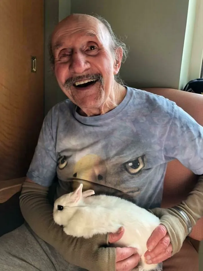 Hombre mayor sonriente sentado con un conejo blanco en brazos, mostrando animales adorables para alegrarte el día.