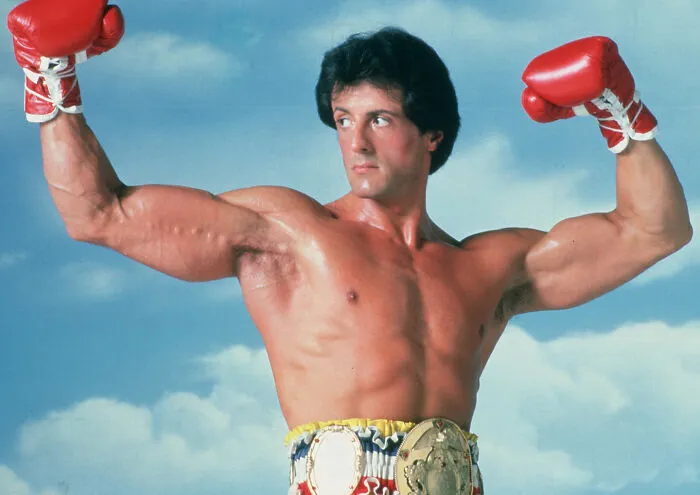 Boxer wearing red gloves and championship belts flexing muscles outdoors in iconic sports pose, interesting facts not learned at school