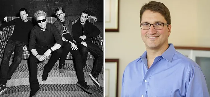 Black and white photo of a band sitting on a couch next to a man in glasses and a blue shirt smiling indoors, interesting facts.