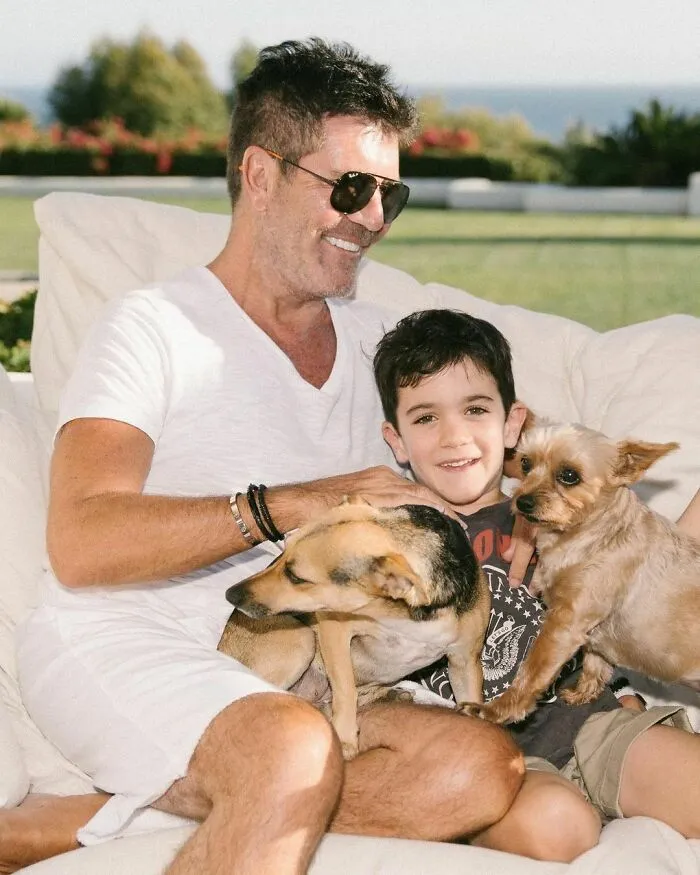 Man and child smiling outdoors, cuddling two dogs, showing immense love for dogs in a relaxed and happy moment.