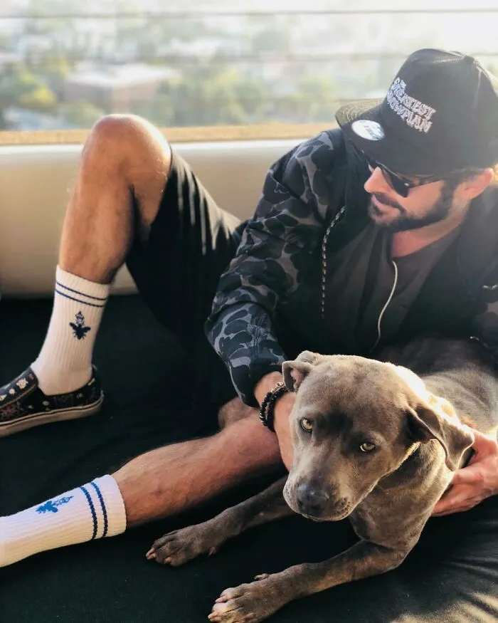Man wearing sunglasses and cap relaxing with a gray dog, showing a celebrity's love for dogs in a casual setting.