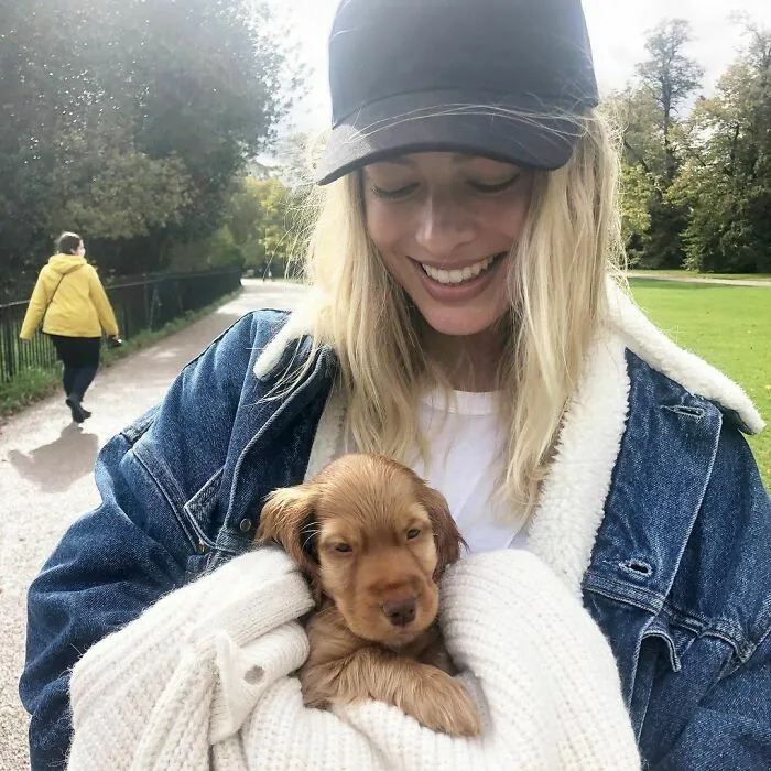 Smiling woman wearing a black cap and blue jacket holding a small brown puppy, showing immense love for dogs outdoors.