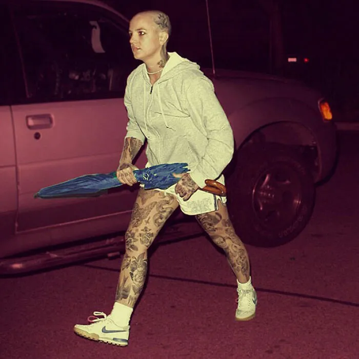 Person with tattoos holding an umbrella, wearing a hoodie and sneakers, stands by a car at night.