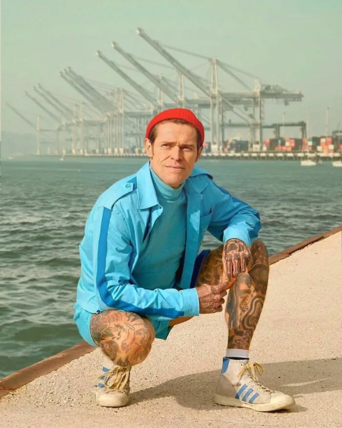 Person in blue outfit and red beanie, tattooed, crouching by the water with cranes in the background.
