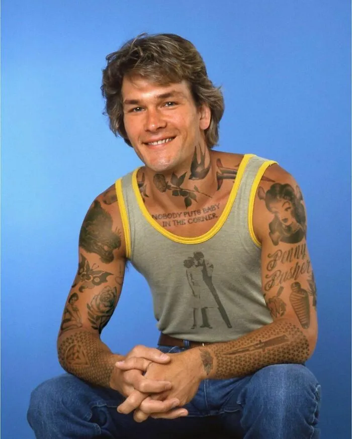 Man with photoshopped tattoos sits wearing a tank top and jeans, showcasing body art on arms and neck.