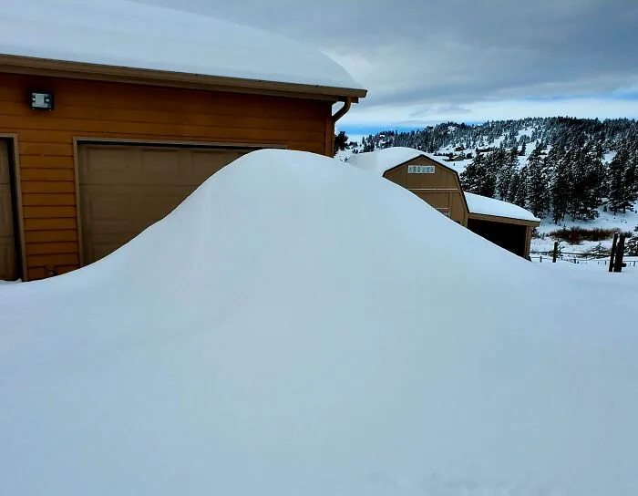 Large snowdrift covering driveway near a garage with snowy mountains and trees in the background showcasing nature challenges.