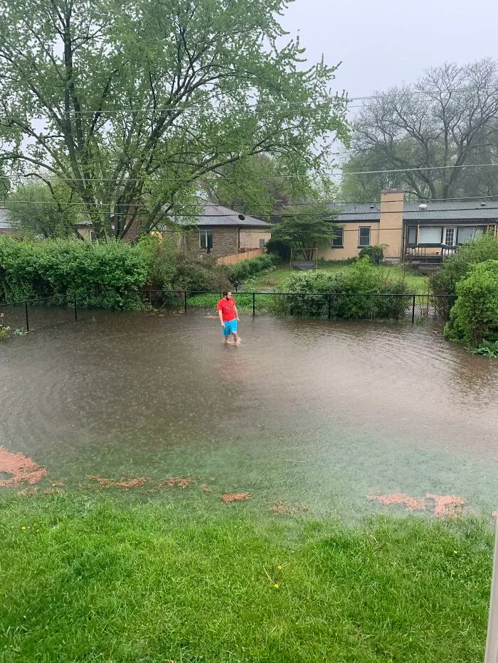 Person standing in a flooded backyard with water reaching mid-calf during heavy rain, showing nature's impact on people.