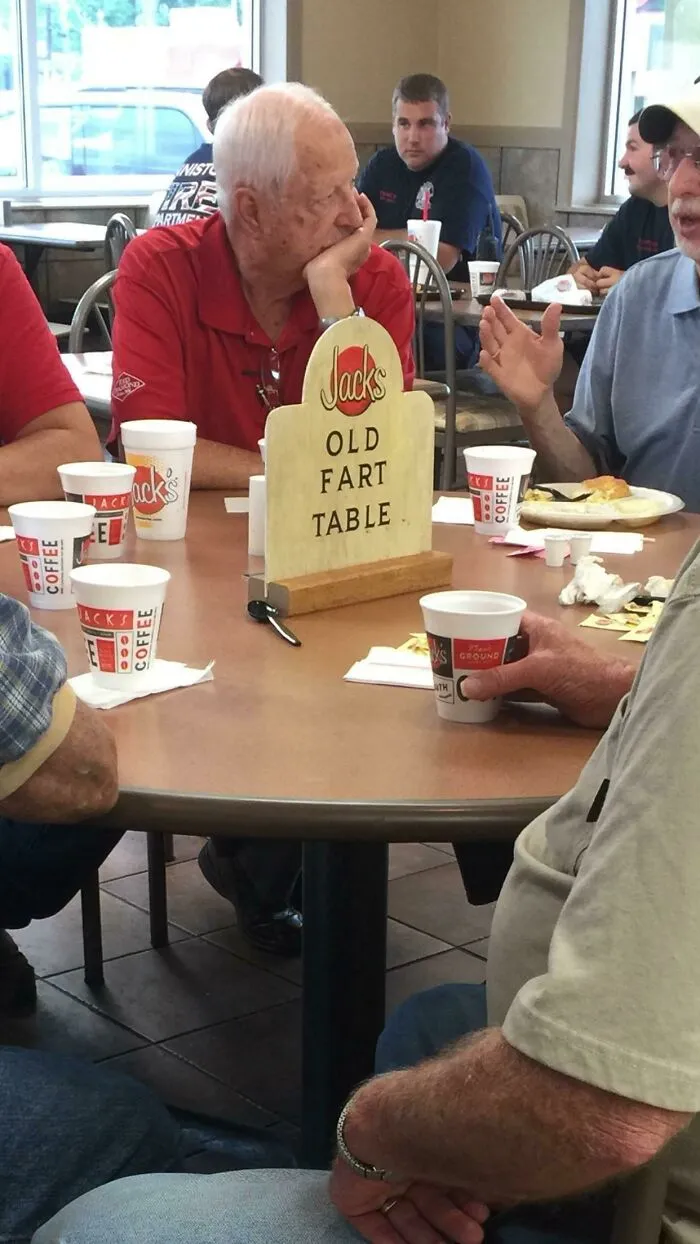 A humorous sign on a table labeled "Old Fart Table" with elderly men sitting around in a fast food restaurant.