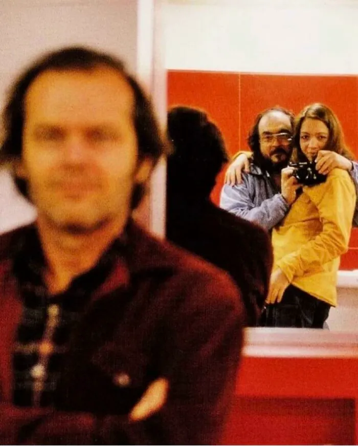 Behind the scenes capture of a historical film set with blurred figure in foreground and couple in mirror reflection.