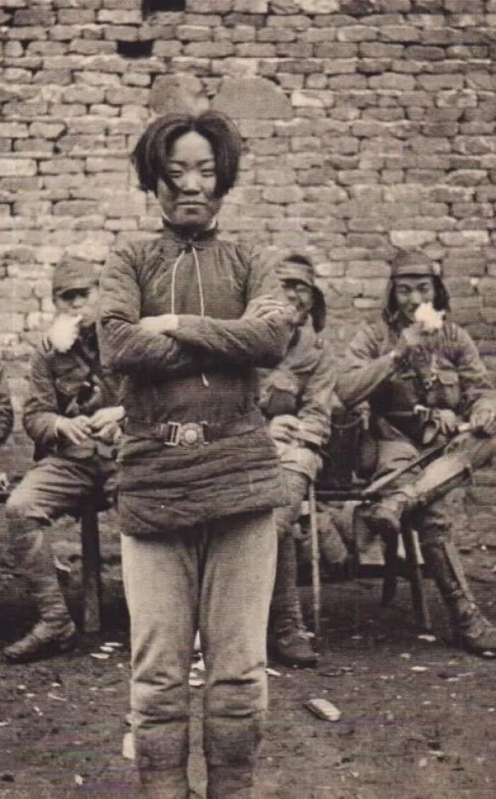 Historical photo of a woman in uniform standing with crossed arms, soldiers seated behind her against a brick wall.